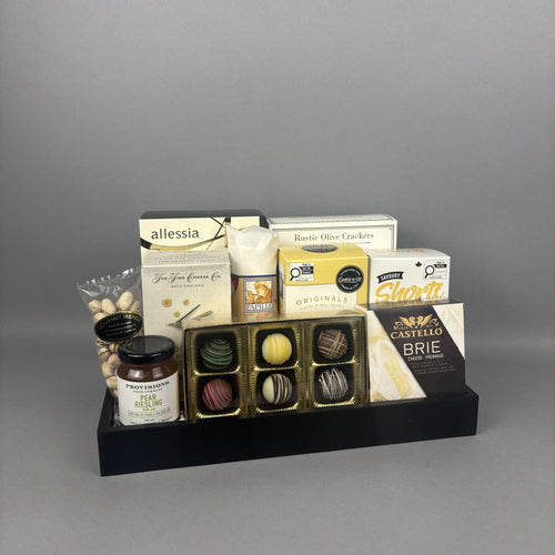 Gourmet sweet and savoury gift basket featuring Castello Brie cheese, Provisions jam, handcrafted chocolate truffles, and assorted gourmet treats arranged on a black rectangular tray.