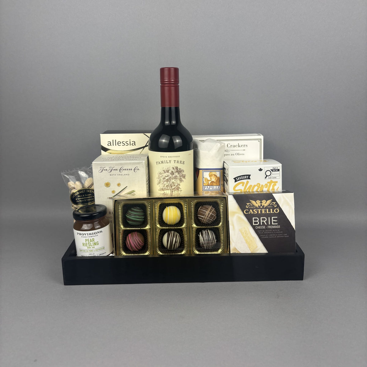 A gourmet gift hamper arranged on a black rectangular tray. The set includes a bottle of red wine, assorted chocolates in a gold tray, a box of Castello Brie cheese, a box of truffles, crackers, cookies, and jam.