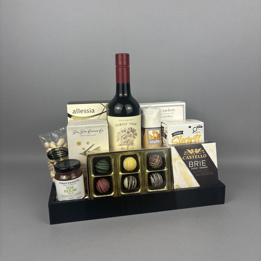 A gourmet gift tray arranged on a black tray, featuring a bottle of red wine, assorted chocolate boxes, a jar of Provisions jam,  chocolates, and other packaged food items.
