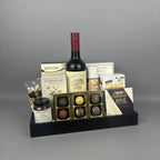 A gourmet gift tray arranged on a black tray, featuring a bottle of red wine, assorted chocolate boxes, a jar of Provisions jam,  chocolates, and other packaged food items.