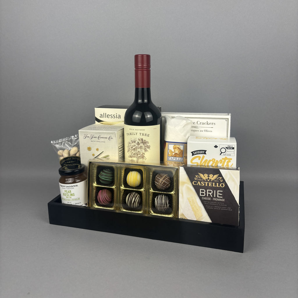 A gourmet gift tray set on a black tray, featuring a bottle of red wine, a box of assorted chocolates, Castello Brie cheese, Allessia cookies, and other gourmet treats.