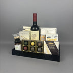 A gourmet gift tray set on a black tray, featuring a bottle of red wine, a box of assorted chocolates, Castello Brie cheese, Allessia cookies, and other gourmet treats.