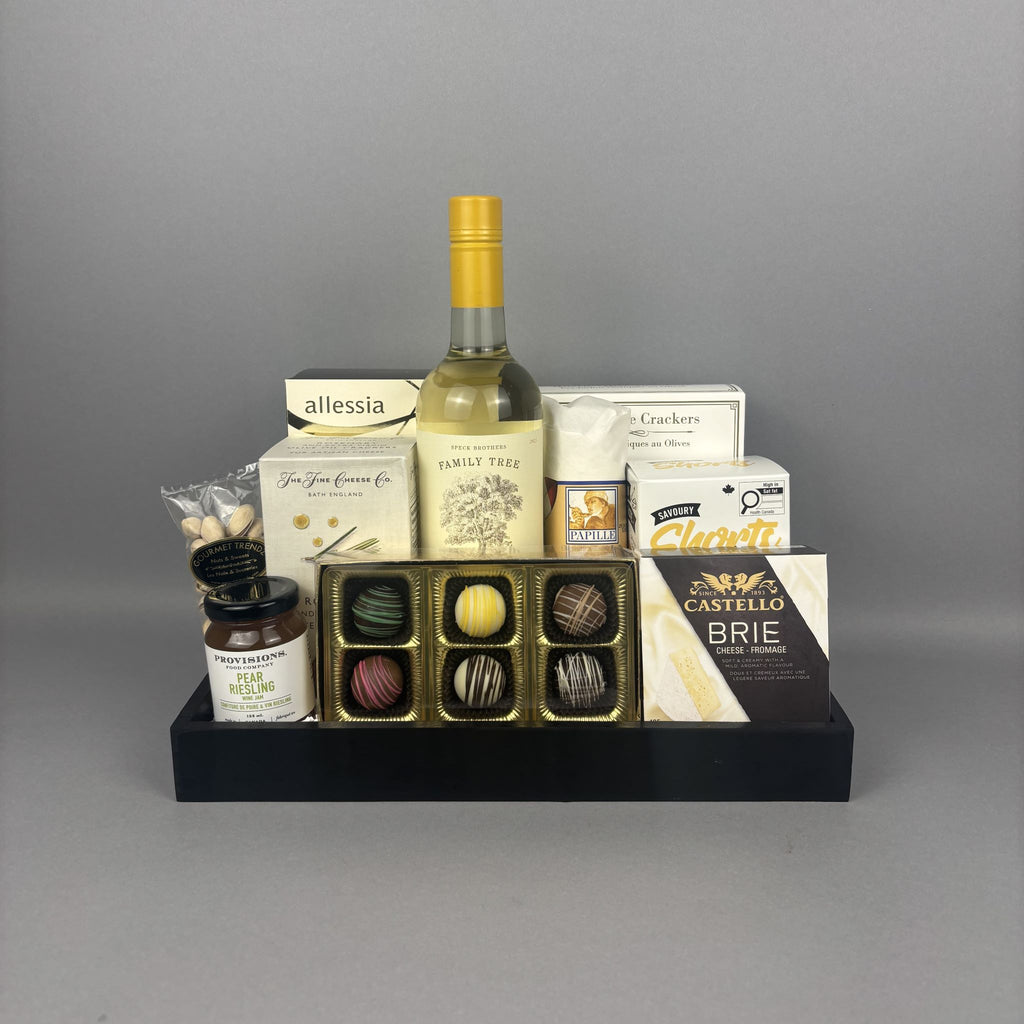 A gourmet gift hamper arranged on a black tray, featuring a bottle of white wine, assorted chocolates, crackers, cheese, and small jars.