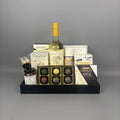 A gourmet gift hamper arranged on a black tray, featuring a bottle of white wine, assorted chocolates, crackers, cheese, and small jars.