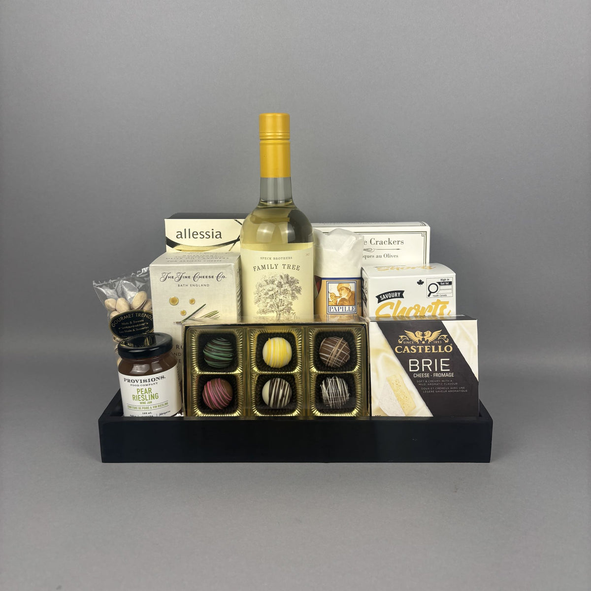 A gourmet gift hamper arranged on a black tray, featuring a bottle of white wine, assorted chocolates, crackers, cheese, and small jars.