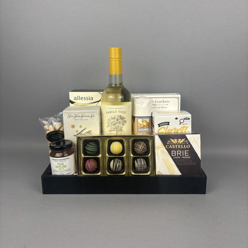 A gourmet gift hamper arranged on a black tray, featuring a bottle of white wine, assorted chocolates, crackers, cheese, and small jars.
