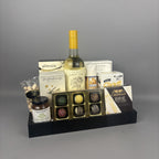 A gourmet gift hamper arranged in a black tray. The hamper includes a bottle of white wine, assorted chocolates, brie cheese, crackers, nuts, and a jar of preserves.