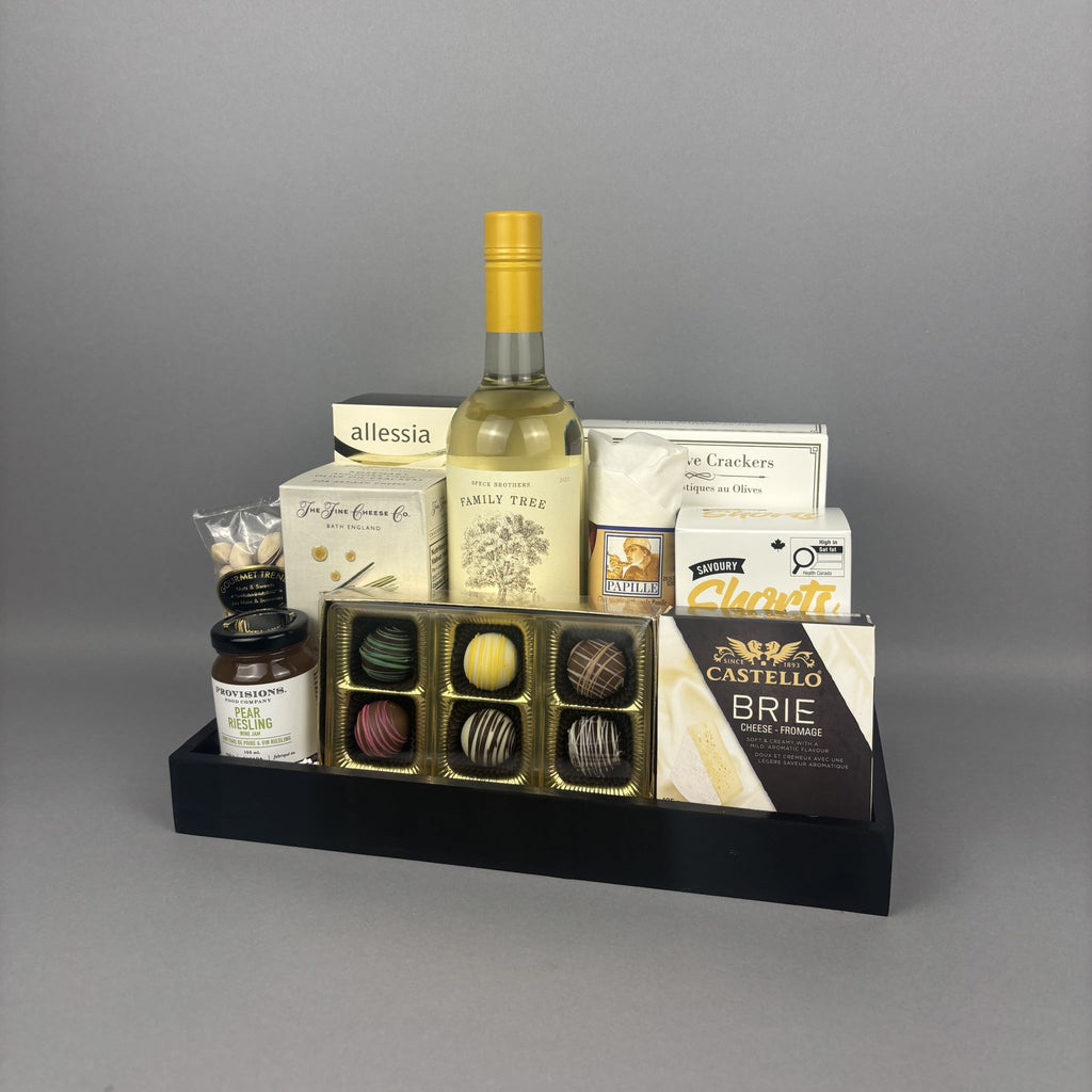 A gourmet gift hamper arranged on a black tray. The set includes a bottle of white wine, assorted chocolates in a gold tray, a box of Castello Brie cheese, salami, and other packaged food items.