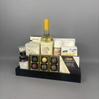 A gourmet gift hamper arranged on a black tray. The set includes a bottle of white wine, assorted chocolates in a gold tray, a box of Castello Brie cheese, salami, and other packaged food items.