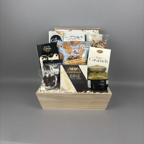 A gourmet gift basket containing a jar of artichoke bruschetta, a box of Castello Brie cheese, cookies, pretzels, and chocolate, arranged in a light wood box.