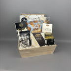 A wooden gift basket filled with assorted gourmet snacks and sweets, including Castello Brie cheese, Caramels, and other packaged treats.