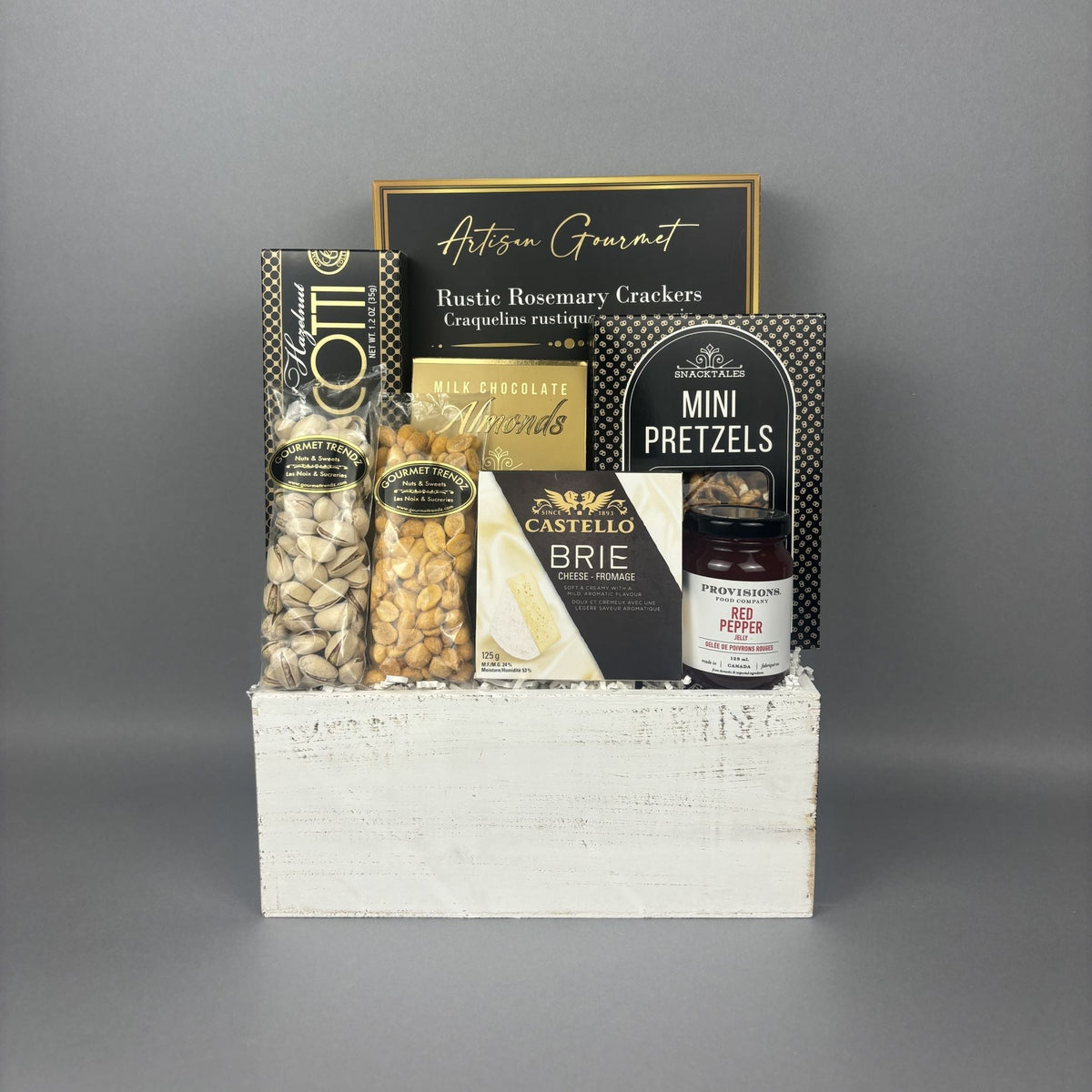 A gourmet gift basket arranged in a white wooden crate. The basket contains a box of almond hazelnut biscotti, pistachios, bbq peanuts, a box of Castello Brie cheese, a box of crackers, a box of mini pretzels, and a jar of Provisions red pepper jelly.