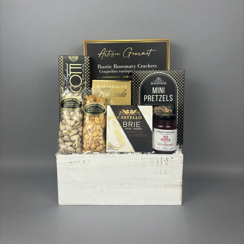 A gourmet gift basket arranged in a white wooden crate. The basket contains a box of almond hazelnut biscotti, pistachios, bbq peanuts, a box of Castello Brie cheese, a box of crackers, a box of mini pretzels, and a jar of Provisions red pepper jelly.