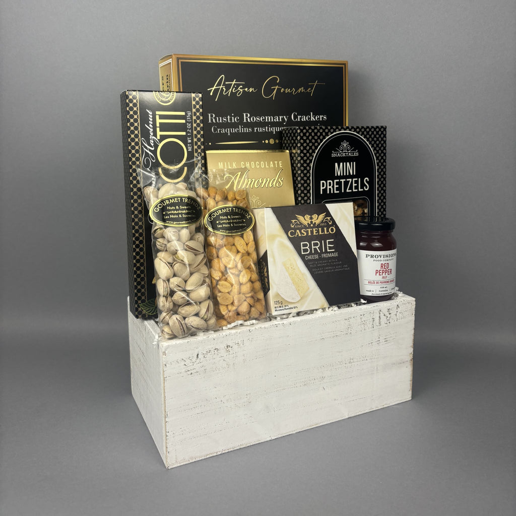 A gourmet gift basket of snacks and cheeses arranged in a distressed white wooden crate. The basket includes a box of Artisan Gourmet crackers, a bag of pistachios, milk chocolate almonds, Castello Brie cheese, mini pretzels, and a jar of Provisions red pepper jelly.