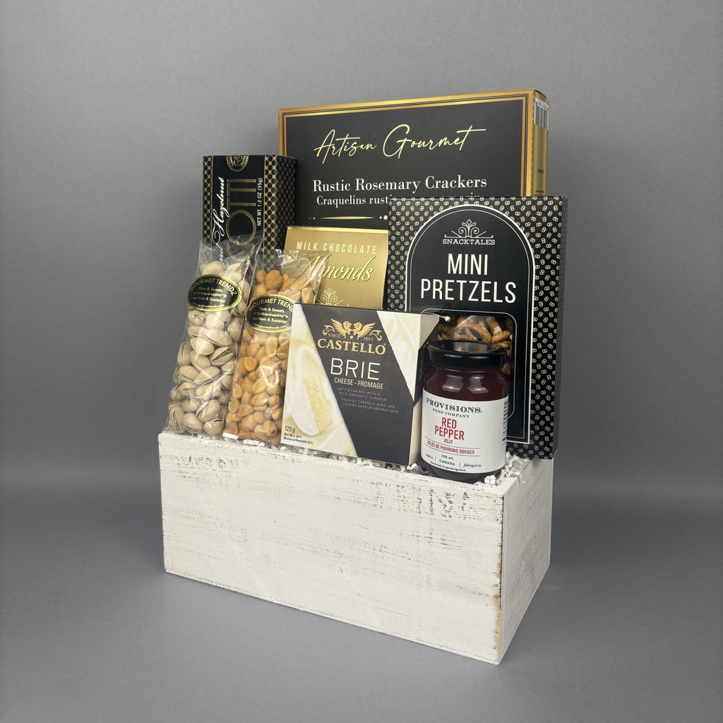A gourmet gift basket containing assorted snacks and cheeses, presented in a distressed white wooden crate. The basket includes Castello Brie cheese, mini pretzels, Rosemary Crackers, and a jar of Red Pepper jelly.