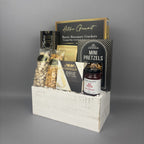 A gourmet gift basket containing assorted snacks and cheeses, presented in a distressed white wooden crate. The basket includes Castello Brie cheese, mini pretzels, Rosemary Crackers, and a jar of Red Pepper jelly.