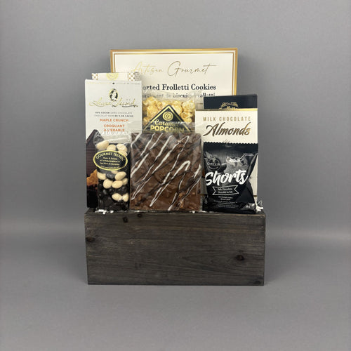 A gourmet chocolate and snack gift basket arranged in a dark rustic wooden box. The basket contains assorted packaged snacks including milk chocolate bar, caramel popcorn, milk chocolate almonds, milk chocolate shortbread, and gourmet frolletti cookies.