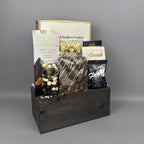 A gourmet chocolate and snack gift basket arranged in a dark wooden box. The basket contains assorted packaged snacks including chocolate chip cookies, milk chocolate almonds, shortbread, and a box of gourmet biscotti.