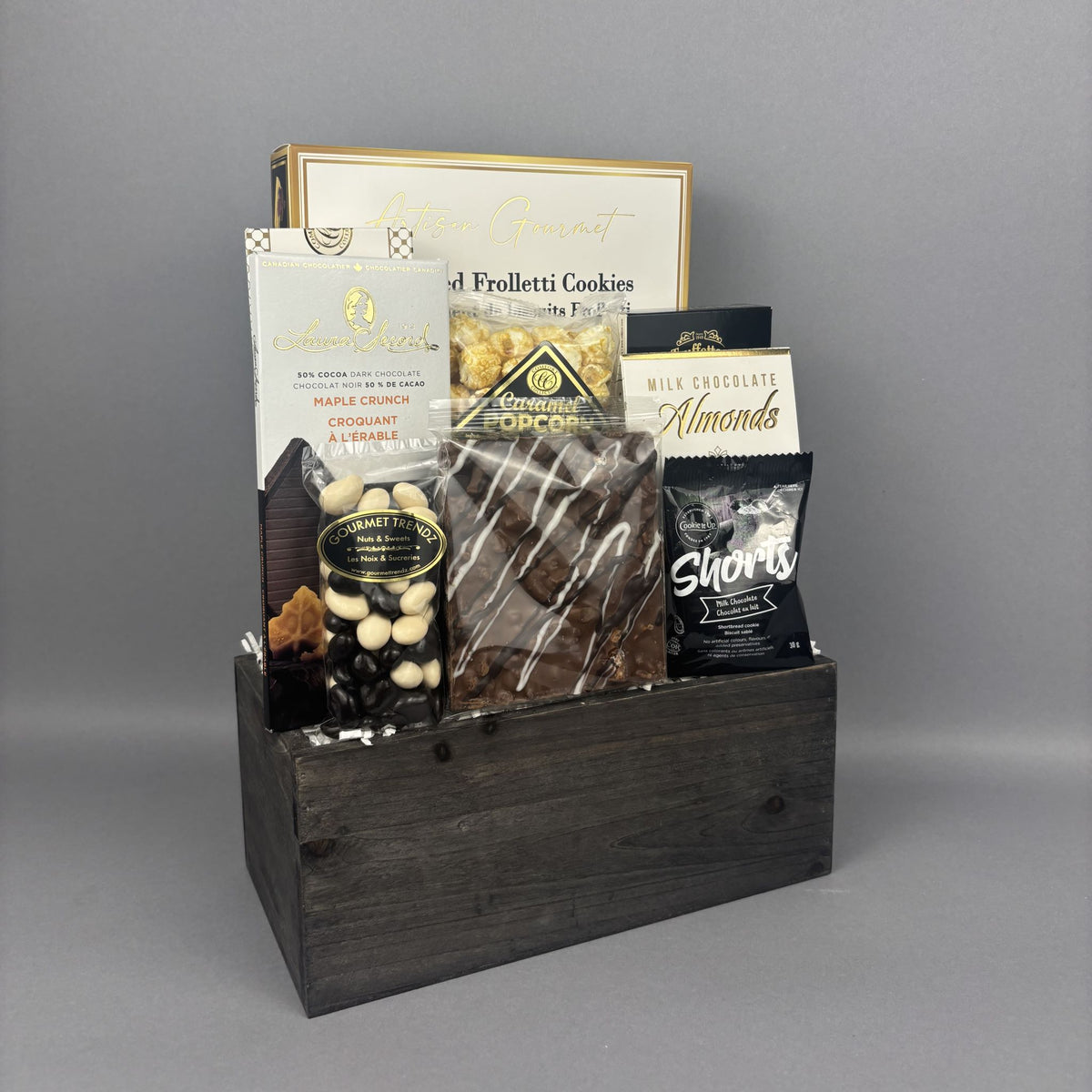A gourmet chocolate and snack gift basket arranged in a dark wooden box. The basket contains assorted packaged snacks including chocolate chip cookies, milk chocolate almonds, shortbread, and a box of gourmet biscotti.