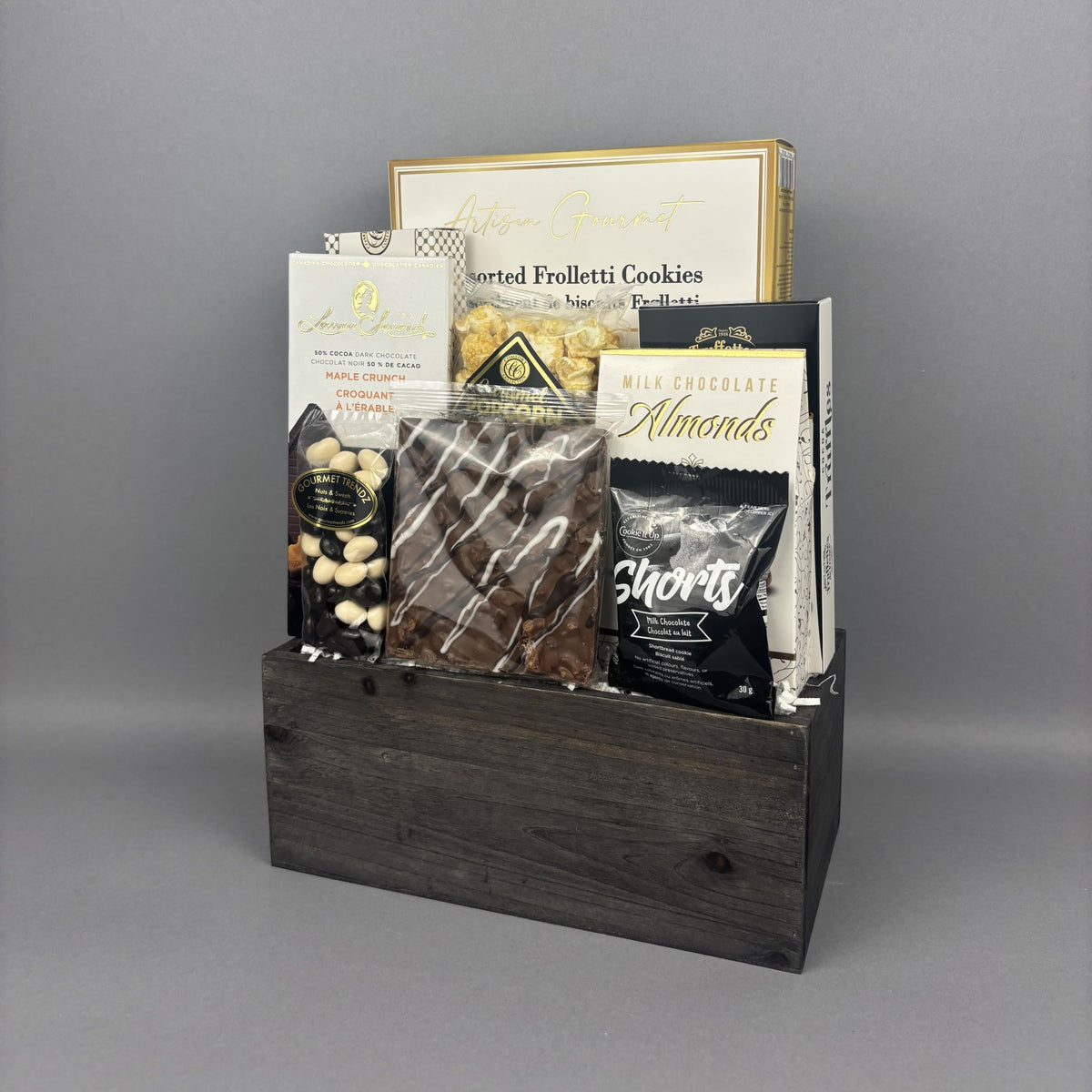 A gourmet gift basket containing assorted chocolates, cookies, and snacks arranged in a dark wooden crate.