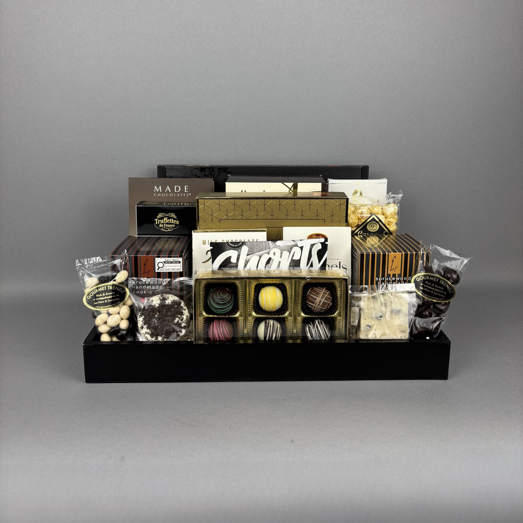 A chocolate gift basket display on a black serving tray, featuring assorted chocolates, truffles, packaged sweets, and brands including Made, Cookie It Up, Hamlet, Lexus, and more.