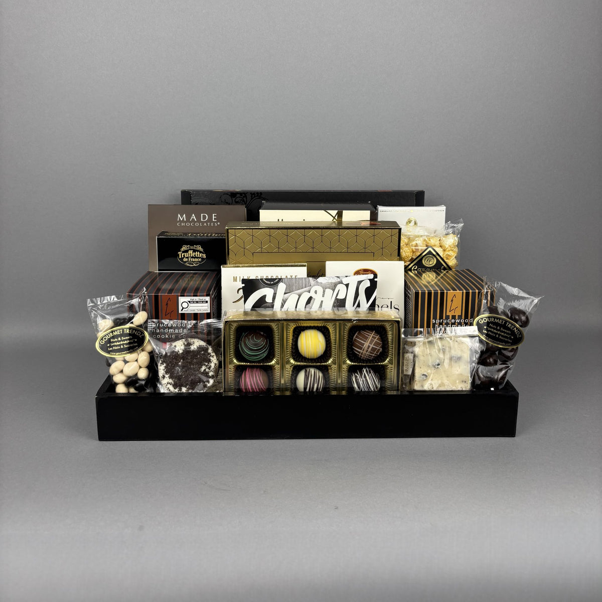 A chocolate gift basket display on a black serving tray, featuring assorted chocolates, truffles, packaged sweets, and brands including Made, Cookie It Up, Hamlet, Lexus, and more.