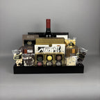 A luxury chocolate and wine gift basket arranged in a black wooden tray. The basket includes a bottle of red wine, assorted gourmet chocolates, truffles, shortbread cookies, and packaged sweets from brands like Made Chocolate and Hamlet.
