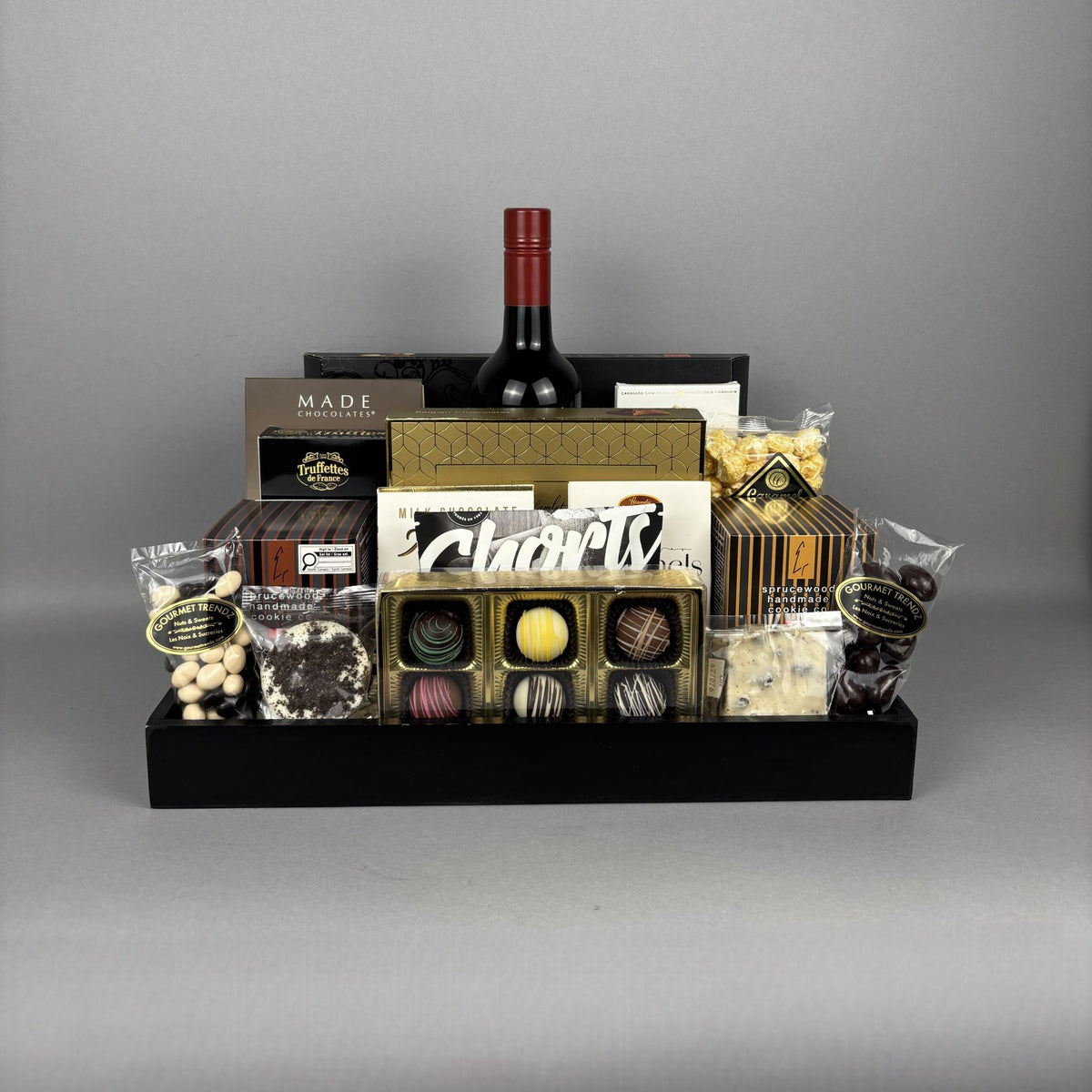 A luxury chocolate and wine gift basket arranged in a black wooden tray. The basket includes a bottle of red wine, assorted gourmet chocolates, truffles, shortbread cookies, and packaged sweets from brands like Made Chocolate and Hamlet.