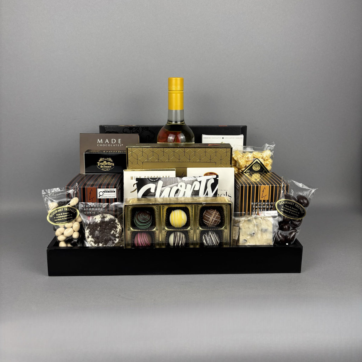 A luxury chocolate and gift basket arrangement in a black rectangular tray, featuring an assortment of chocolates, truffles, packaged sweets, a bottle of white wine.