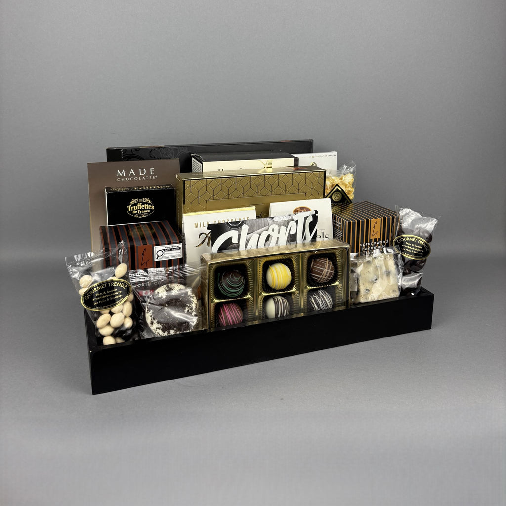A luxury chocolate gift hamper arranged on a black rectangular tray. The assortment includes boxed chocolates, pralines, and packaged sweets from brands such as Made Chocolates and Laura Secord.