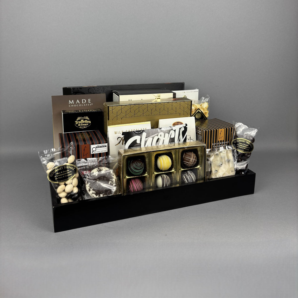 A luxury chocolate gift hamper arranged on a black rectangular tray. The assortment includes boxed chocolates, pralines, and packaged sweets from brands such as Made Chocolates and Laura Secord.