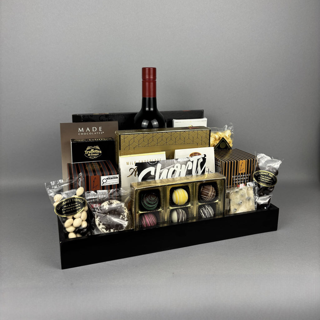 A luxury gourmet gift hamper arranged in a black rectangular tray. The hamper includes a bottle of red wine, assorted chocolates in gold and clear packaging, truffles, packaged nuts, and branded boxes and bags such as Made Chocolate and Sprucewood.