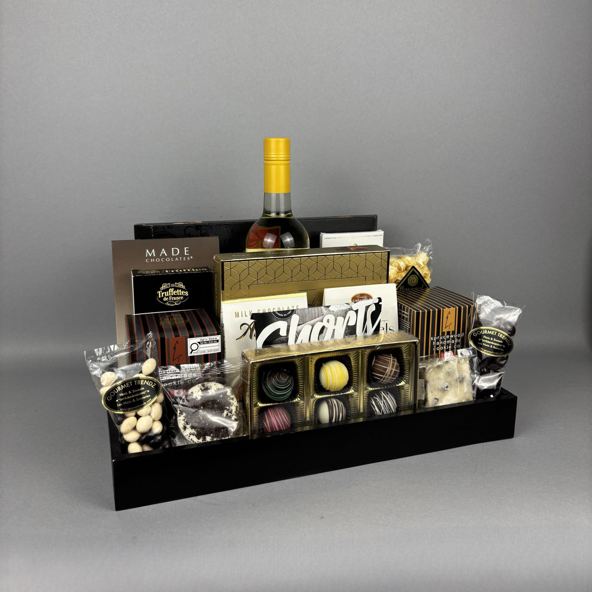 A luxury chocolate and wine gift basket arranged in a black wooden tray. The basket includes a white bottle of wine, assorted chocolates, truffles, shortbread cookies, and packaged sweets from brands like Made Chocolates and Hamlet.