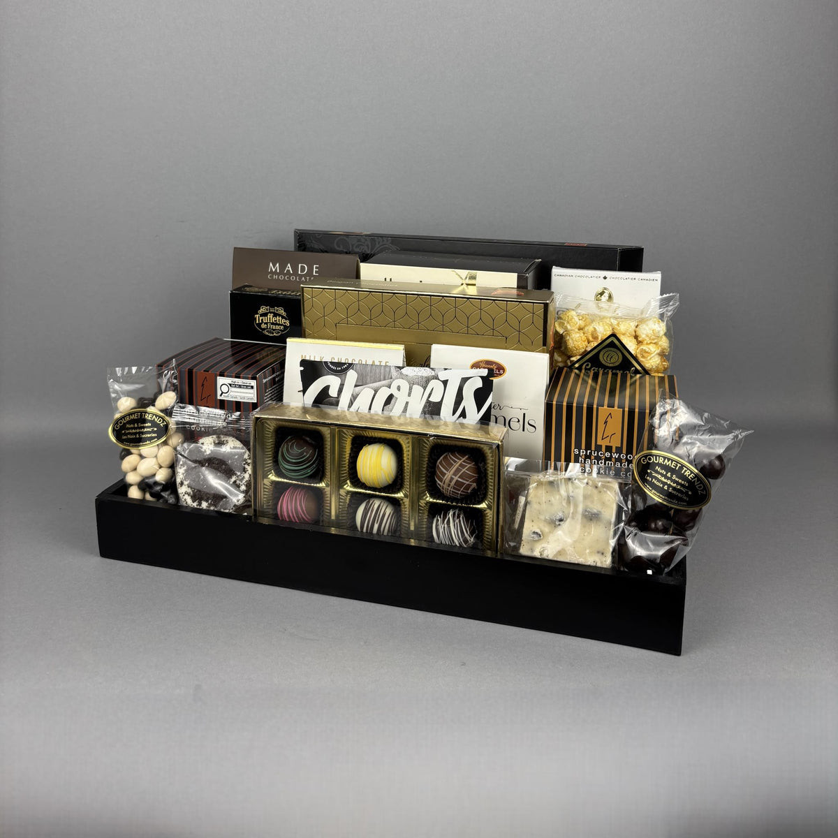 A gourmet chocolate gift basket arranged in a black rectangular tray, featuring assorted chocolates, truffles, fudge, shortbread cookies, and other packaged sweets.
