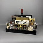 A gourmet gift hamper arranged in a black rectangular tray. The hamper includes a red wine bottle, assorted boxed chocolates, truffles, caramel candies, nuts, and packaged sweets.