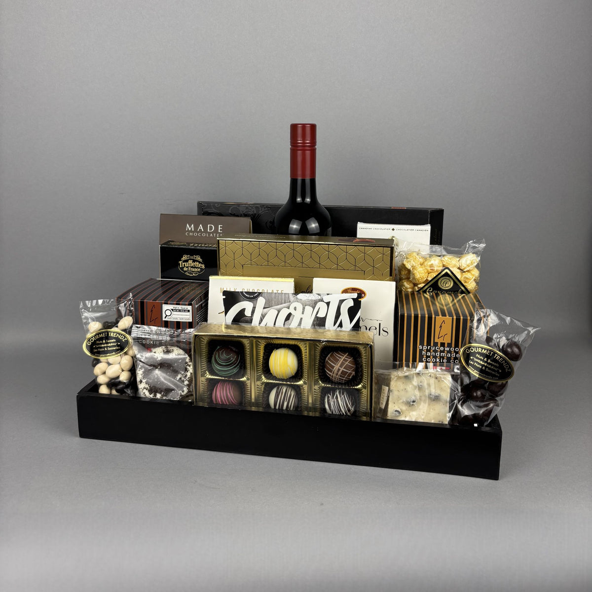 A gourmet gift hamper arranged in a black rectangular tray. The hamper includes a red wine bottle, assorted boxed chocolates, truffles, caramel candies, nuts, and packaged sweets.