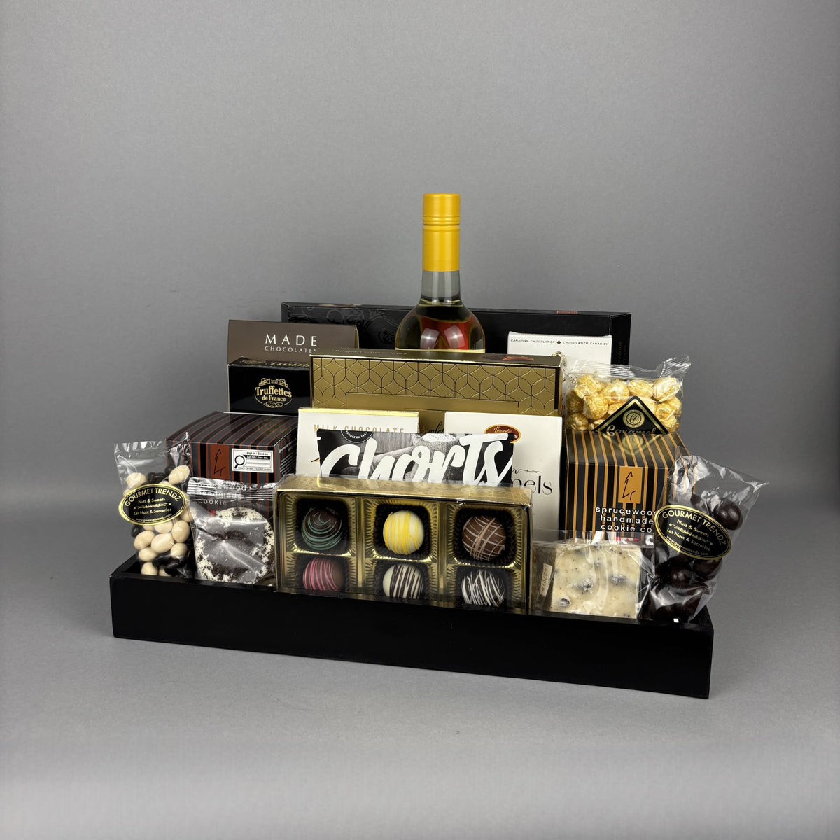 A gourmet chocolate and snack gift basket arranged on a black rectangular tray. The set includes a box of assorted chocolates, several packaged sweets, bags of nuts, a bottle of white wine, and branded boxes.