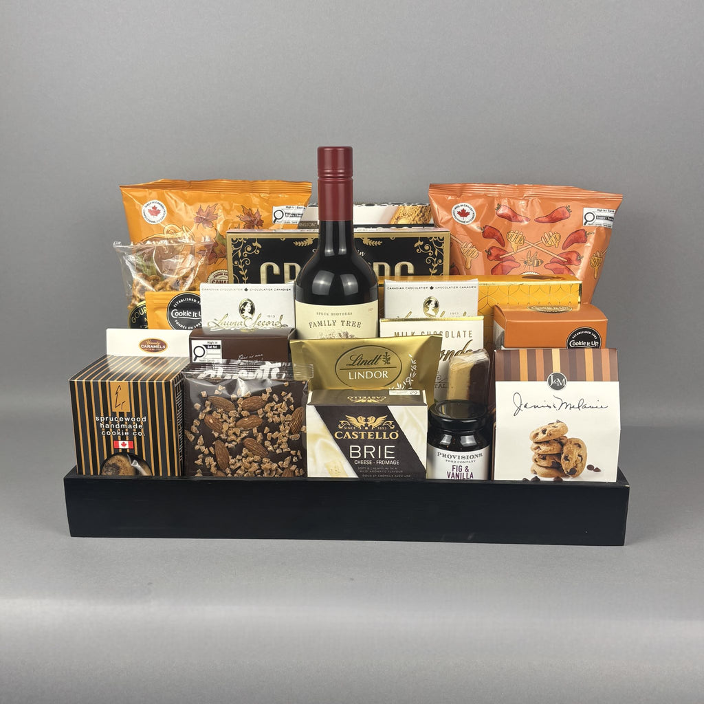 A gourmet gift basket arranged in a black wooden tray, featuring a bottle of red wine, assorted cheeses, crackers, cookies, and packaged snacks.