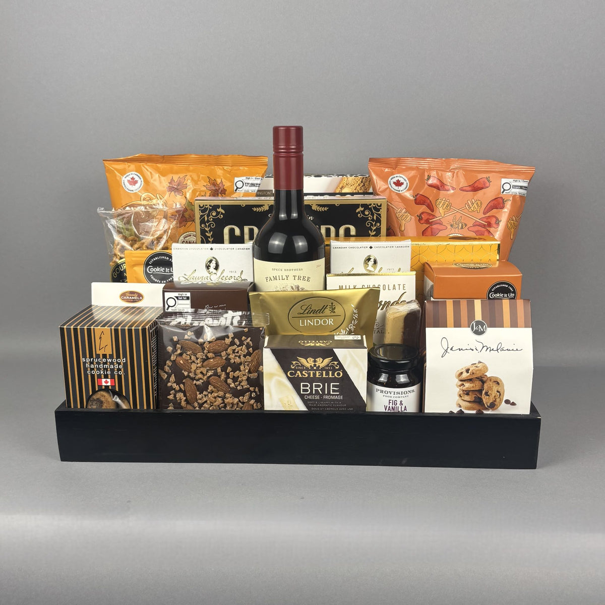 A gourmet gift basket arranged in a black wooden tray, featuring a bottle of red wine, assorted cheeses, crackers, cookies, and packaged snacks.