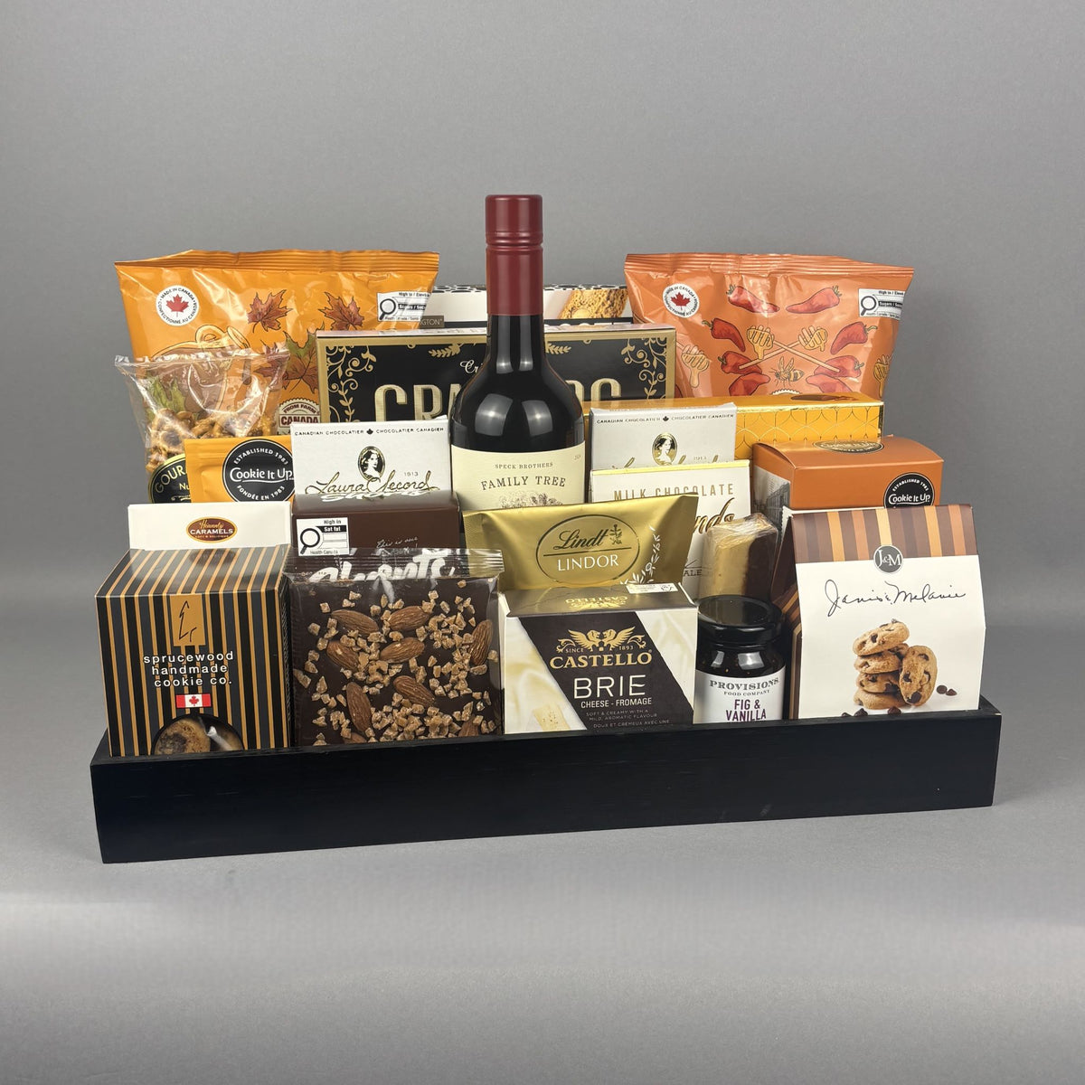 A gourmet gift hamper arranged in a black rectangular tray, featuring a bottle of red wine, assorted cheeses, chocolates, nuts, and packaged snacks.