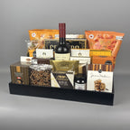 A gourmet gift hamper arranged in a black rectangular tray. The hamper contains a bottle of wine, assorted chocolate boxes, cookies, nuts, and packaged snacks.