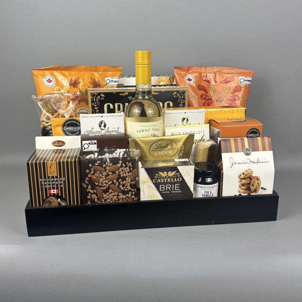A gourmet gift hamper arranged in a black tray, featuring a bottle of white wine, assorted packaged snacks, chocolates, cookies, and a Provisions jam.