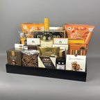 A gourmet gift basket filled with assorted snacks, cheeses, and treats arranged in a black tray. Visible items include a bottle of white wine, a box of Castello Brie, a bag of Sprucewood cookies, and several other packaged food items.