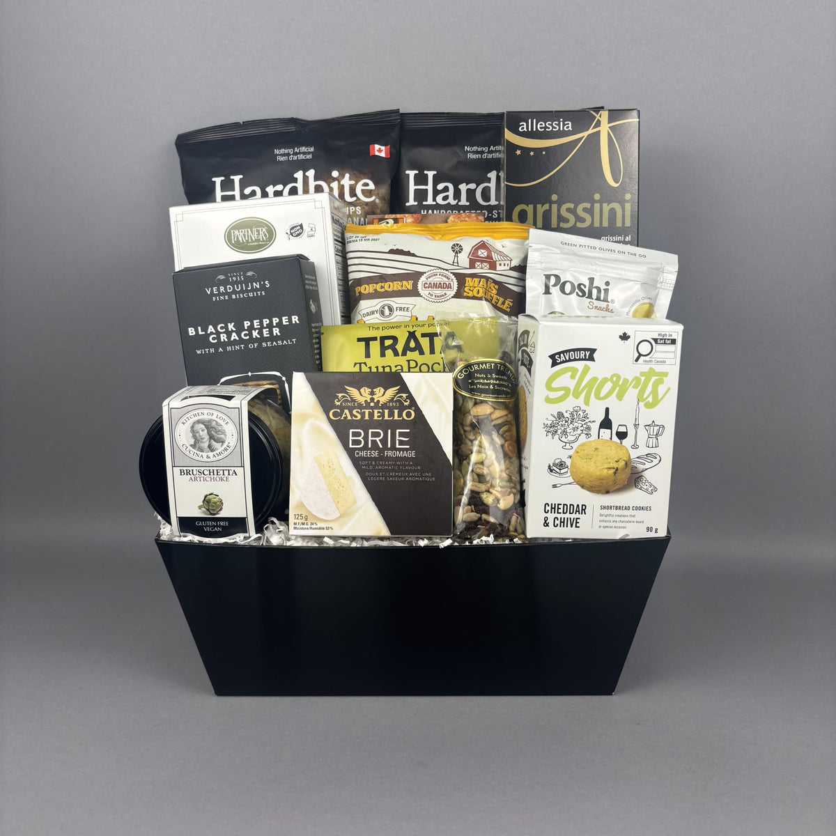 A gourmet gift basket filled with assorted packaged snacks, cheeses, crackers, and artichoke bruschetta, arranged in a black rectangular basket.
