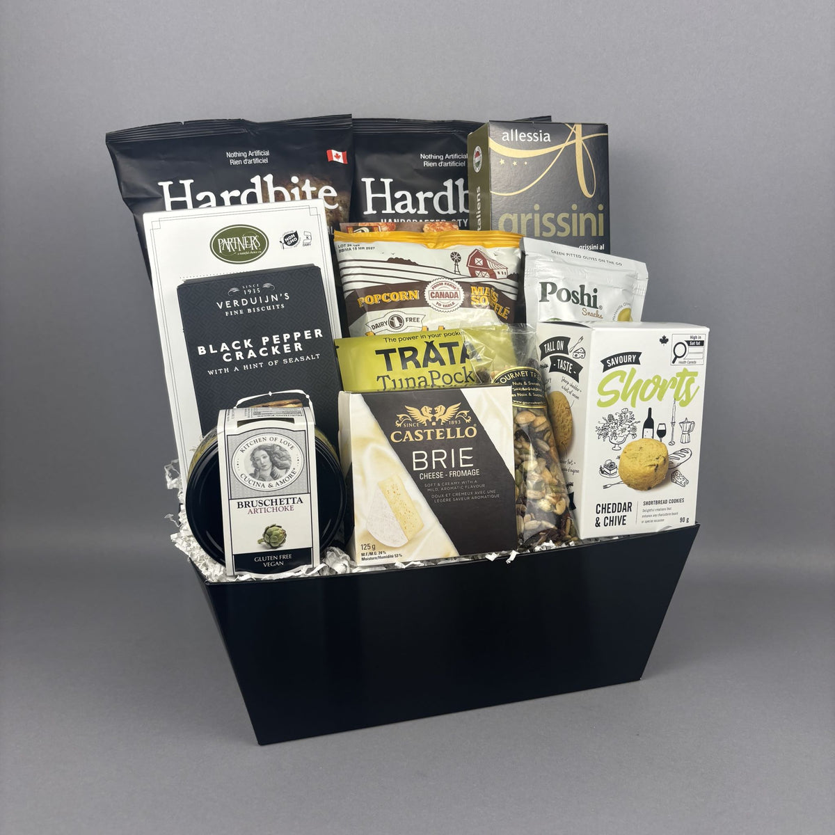A gourmet gift basket filled with assorted snacks and cheeses, presented in a black basket. The basket contains bags of Hardbite handcrafted chips, Poshi green olives, tuna pate, Castello Brie cheese, and other packaged snacks.