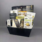 A gourmet gift basket filled with assorted snacks and cheeses, presented in a black rectangular basket. Visible brands include Hardbite, Allessia, Castello, Poshi, and Cookie It Up Shorts.