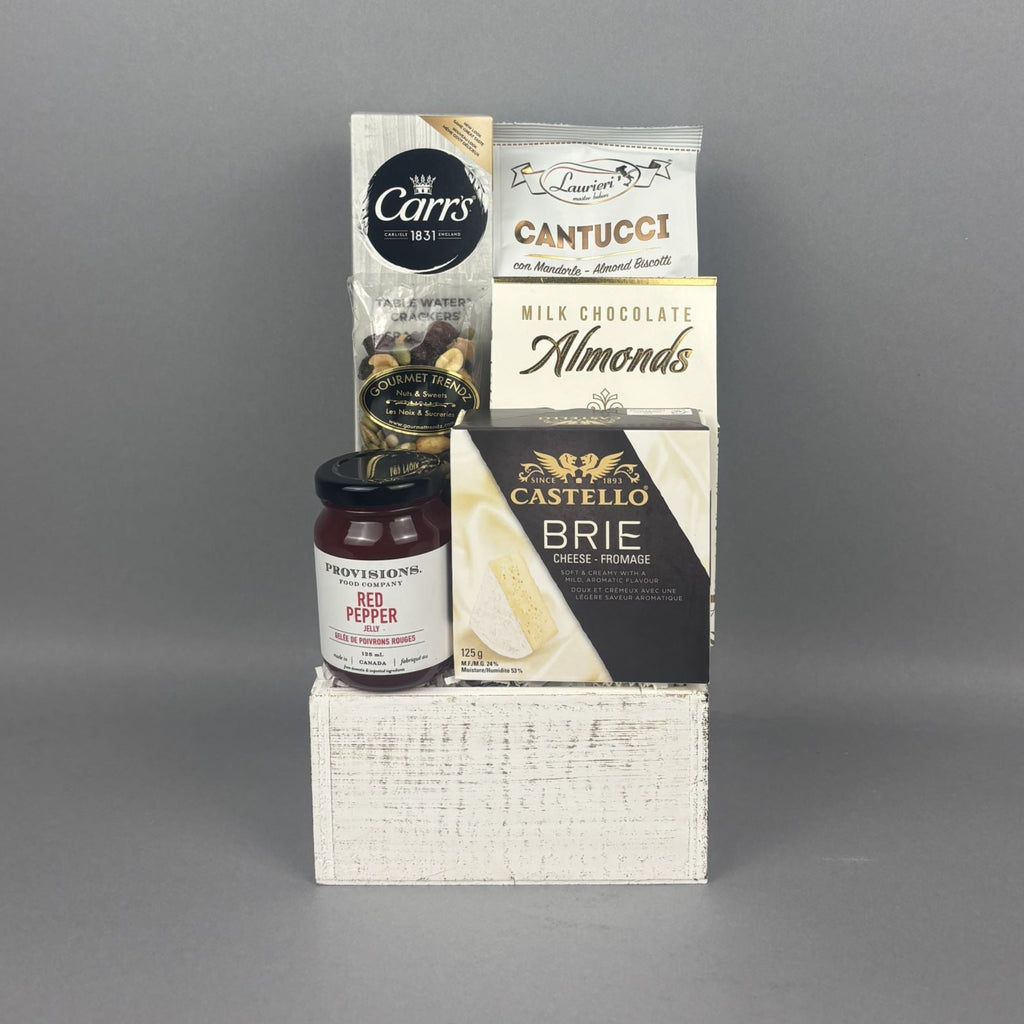 A curated gourmet gift hamper featuring Castello Brie cheese, a box of milk chocolate Almonds, a bag of Cantucci almond biscotti, and a box of Carr's crackers, all arranged on a white rustic wood box.