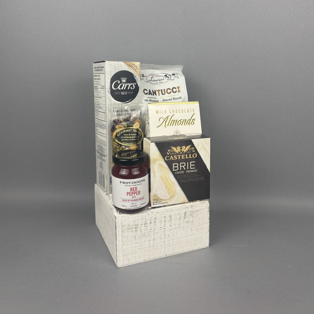 A gift hamper set in a white textured wooden crate, featuring a box of Carr's crackers, Provisions Red Pepper jelly, Cantucci almond biscotti, Milk Chocolate Almonds, Castello Brie cheese.