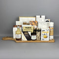 A gourmet gift hamper arranged on a wooden serving board. The assortment includes Trâta Salmon Pocket, Castello Brie, Short's Canadian Cheese Biscuits, a bottle of fresh lime vinegar, pistachios, sea salt breadsticks , and various crackers and sweets.