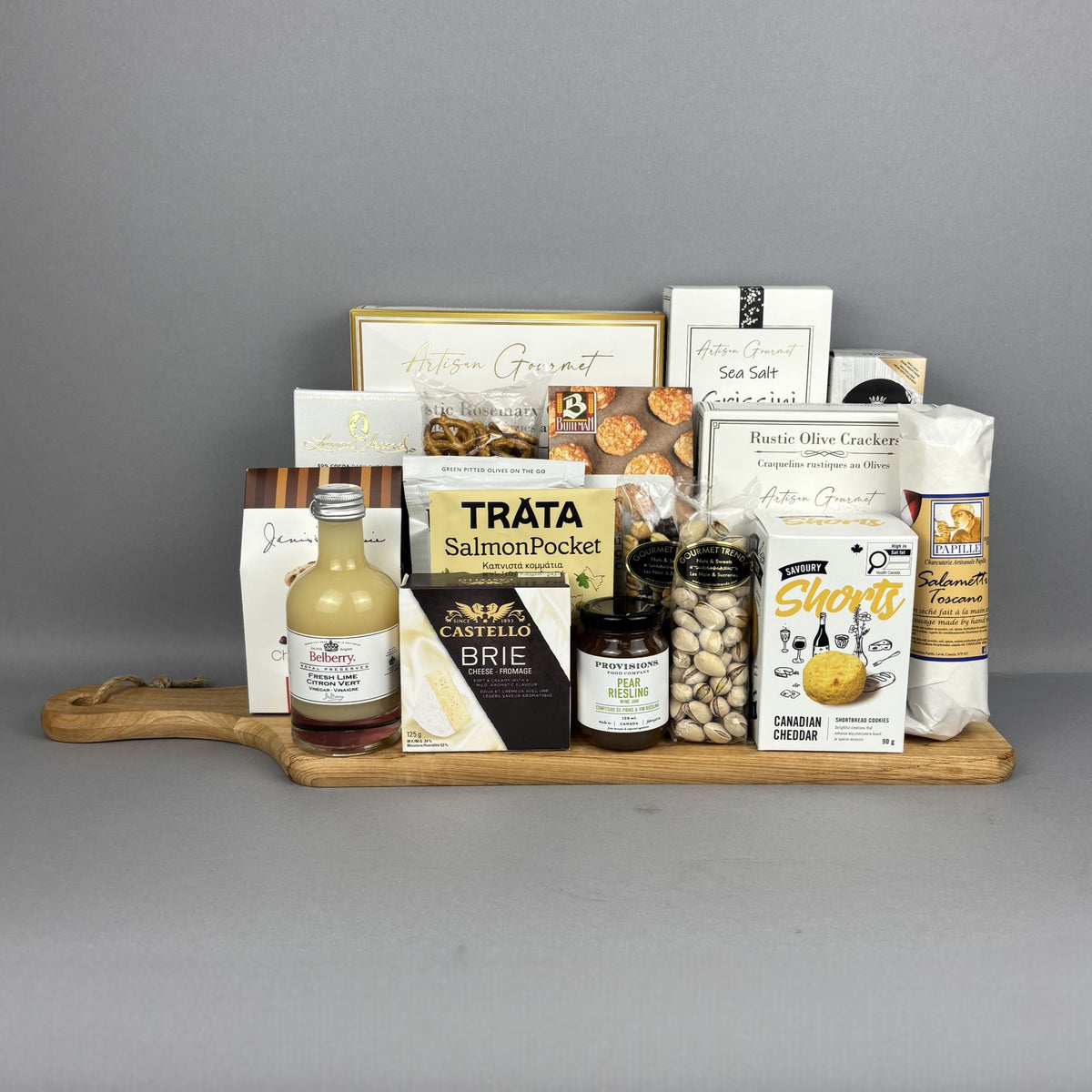 A gourmet gift hamper arranged on a wooden serving board. The assortment includes Trâta Salmon Pocket, Castello Brie, Short's Canadian Cheese Biscuits, a bottle of fresh lime vinegar, pistachios, sea salt breadsticks , and various crackers and sweets.